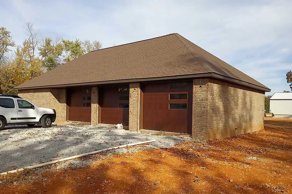 Elite Garage Door Services of Northwest Arkansas-A tan brick building with three dark brown residential garage doors in Rogers, AR, features a gravel driveway and a white SUV parked nearby, showcasing a recent garage door installation by a local professional service in Northwest Arkansas.