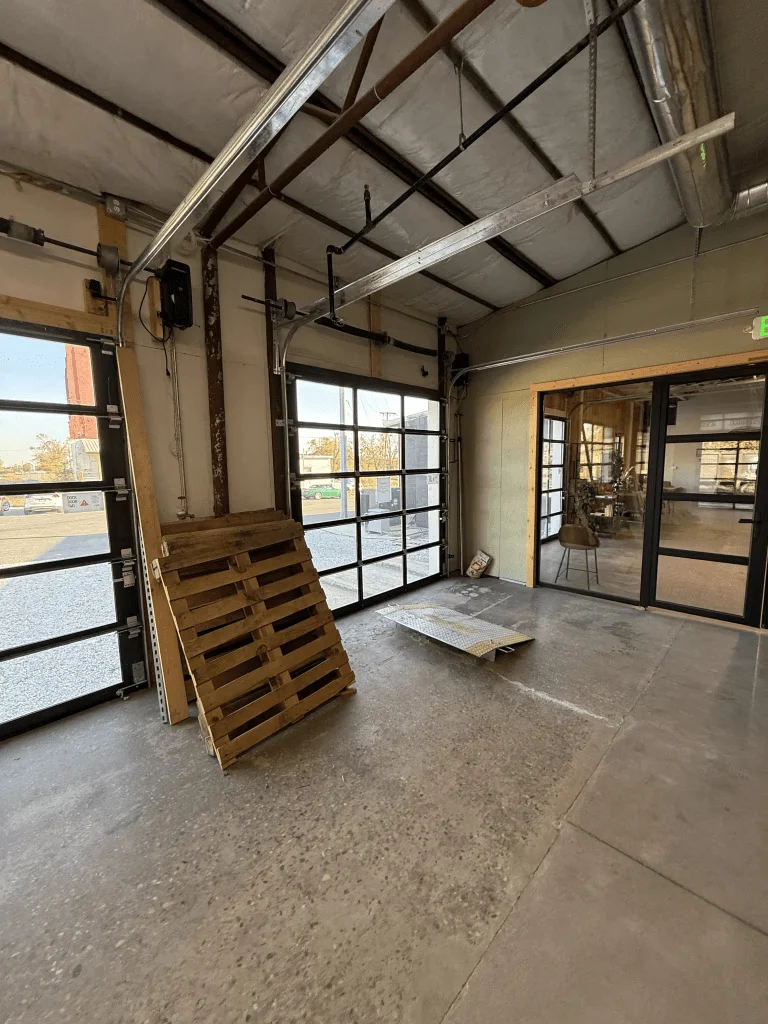 Elite Garage Door Services of Northwest ArkansasThe image shows a spacious garage interior with overhead garage doors partially open, revealing a view outside. A wooden pallet leans against a wall. This scene is typical of a professional garage door service location in Northwest Arkansas.
