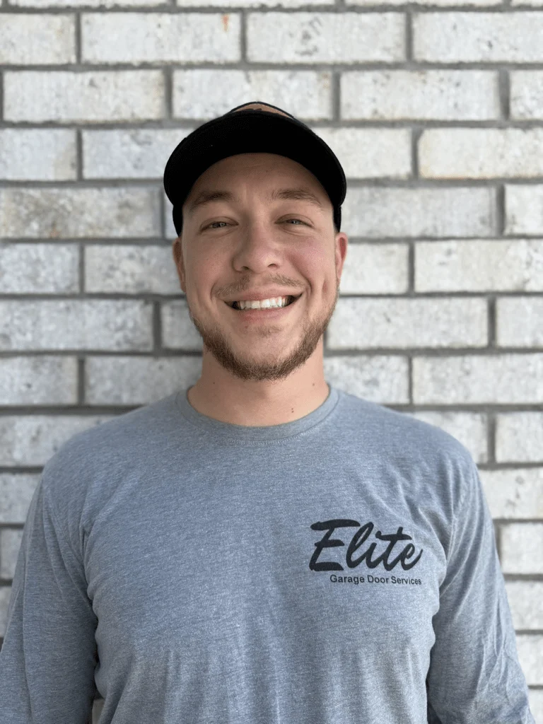 Elite Garage Door Services of Northwest ArkansasA professional from Elite Garage Door Services in Northwest Arkansas, wearing a branded gray shirt and cap, stands smiling against a light brick wall, representing reliable local garage door repair services in NWA.