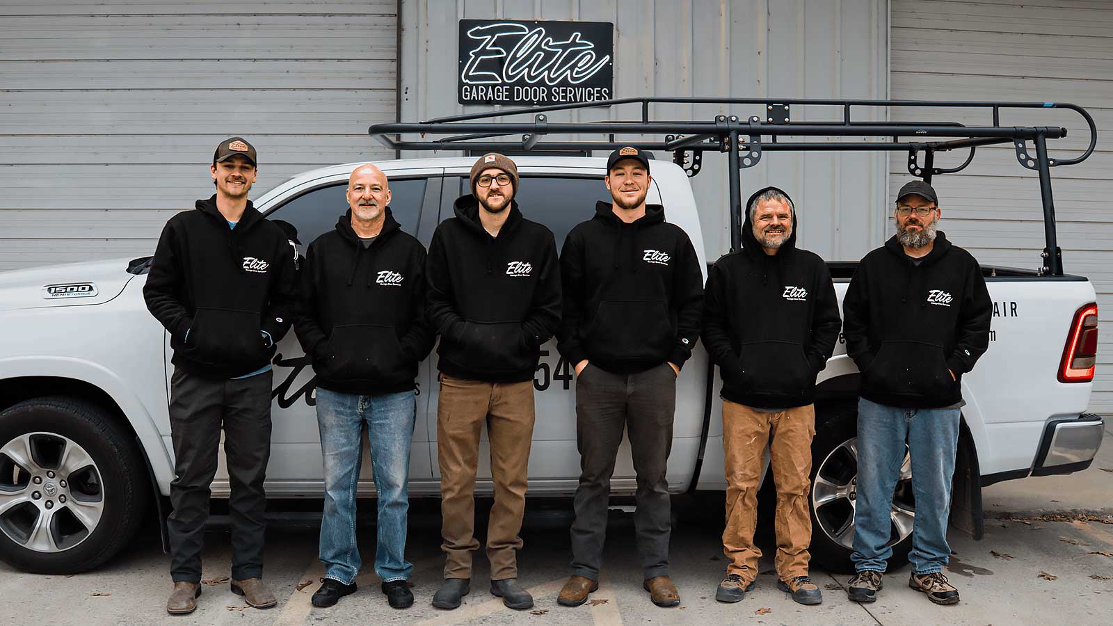 new-homepage-team-image Elite Garage Door Services of Northwest Arkansas-A team of six technicians from Elite Garage Door Services stands in front of a branded truck in Northwest Arkansas, wearing black hoodies, ready to provide reliable garage door repair and installation services.