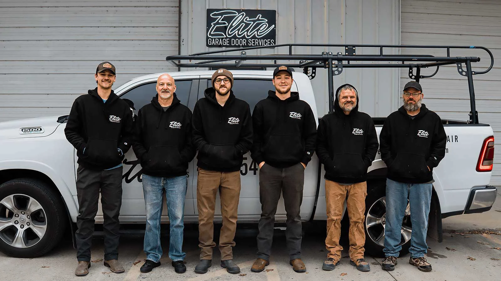 Elite Garage Door Services of Northwest Arkansas-A team of six technicians from Elite Garage Door Services stands in front of a branded truck in Northwest Arkansas, wearing black hoodies, ready to provide reliable garage door repair and installation services.