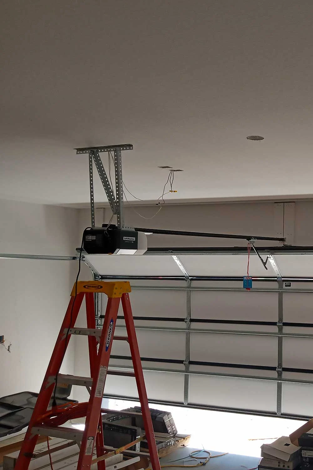 Elite Garage Door Services of Northwest Arkansas-A red ladder is positioned beneath a newly installed garage door opener in a residential garage, highlighting professional garage door installation in Northwest Arkansas. Wires hang from the ceiling, indicating ongoing setup.