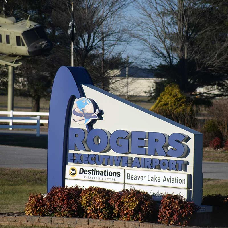 Elite Garage Door Services of Northwest Arkansas-A sign for Rogers Executive Airport stands prominently, surrounded by bushes, with a helicopter in the background. This location in Rogers AR is relevant for local garage door repair and installation services in Northwest Arkansas.
