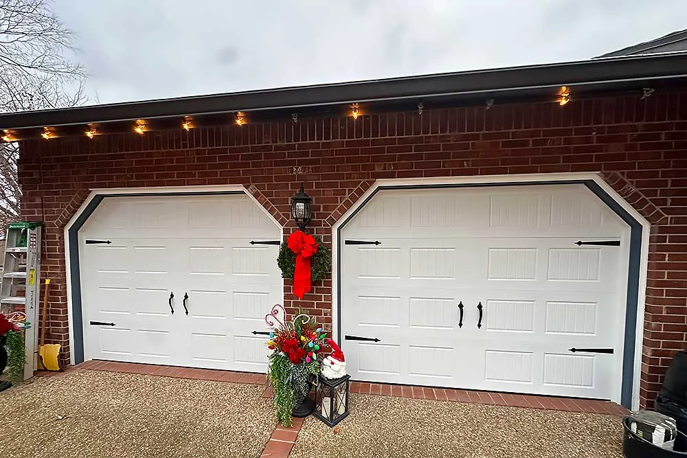 Elite Garage Door Services of Northwest Arkansas-A brick house in NWA features two white residential garage doors adorned with festive wreaths and holiday lights, showcasing professional garage door installation.