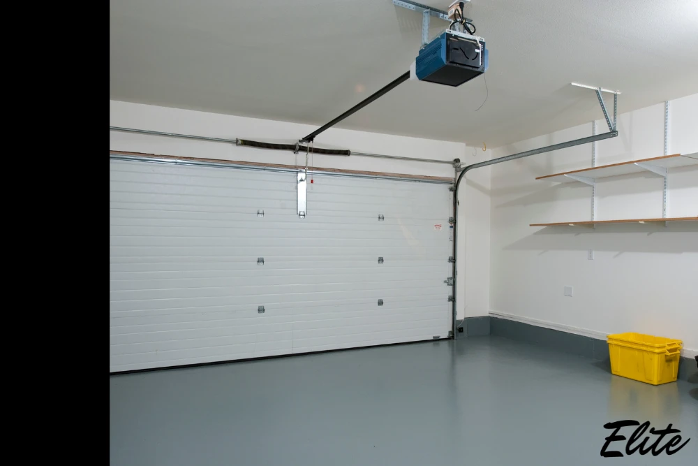 Elite Garage Door Services of Northwest Arkansas-A clean garage interior in Northwest Arkansas features a white overhead garage door with an installed opener, highlighting professional garage door service. Shelves and a yellow storage bin are visible against the white walls.