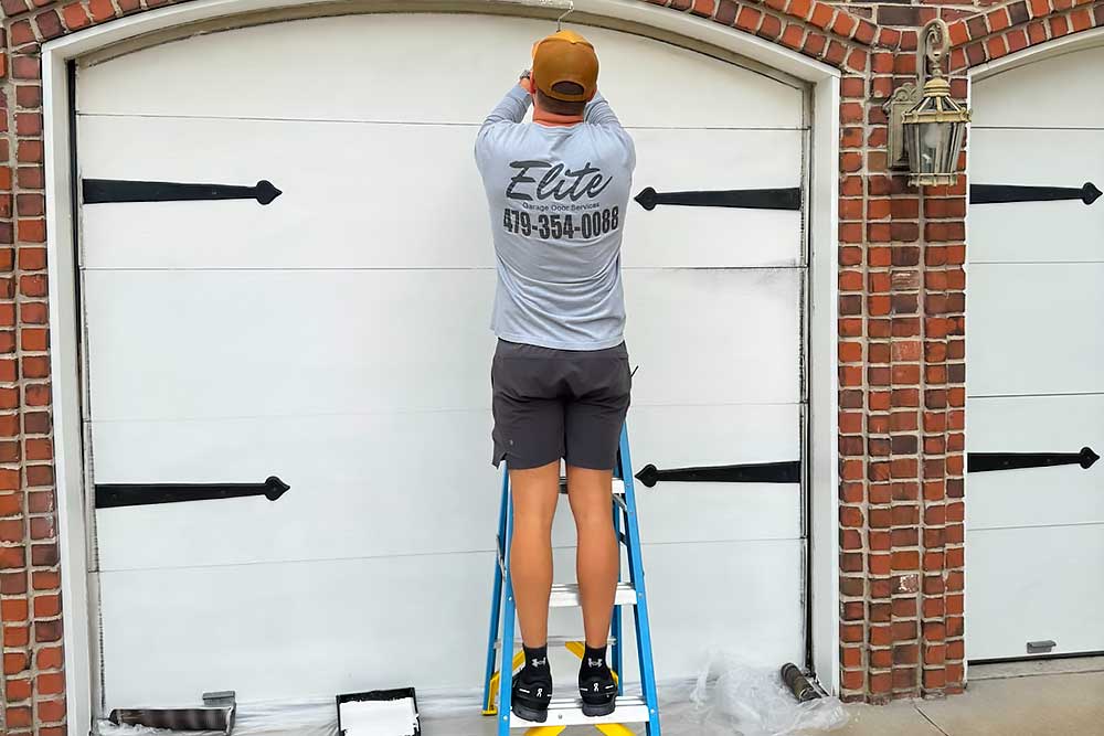 springdale-image Elite Garage Door Services of Northwest Arkansas-A professional technician from Elite Garage Door is performing garage door repair on a white residential garage door in Northwest Arkansas, standing on a ladder against a brick exterior.