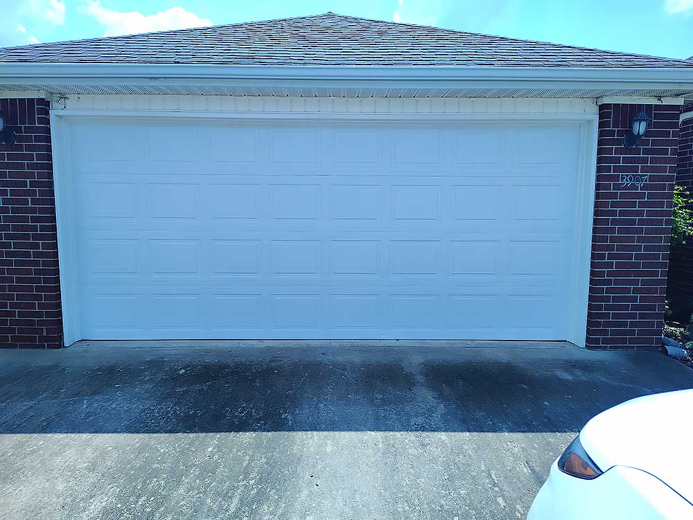 work-carousel-4 Elite Garage Door Services of Northwest Arkansas-A white residential garage door is closed, set in a red brick wall, under a clear blue sky, showcasing a professional installation in Rogers AR. A white car's front end is partially visible in the driveway.
