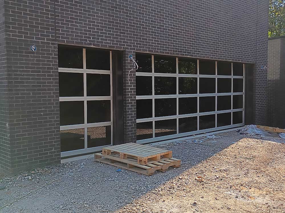 work-carousel-5 Elite Garage Door Services of Northwest Arkansas-Two large, glass-panel residential garage doors are installed on a dark brick building under construction, with wooden pallets on the gravel ground, highlighting professional garage door installation in Northwest Arkansas.