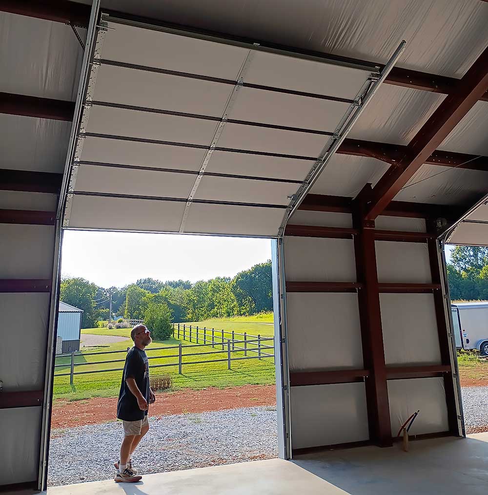 work-carousel-6 Elite Garage Door Services of Northwest Arkansas-A person stands inside a large garage with an open door, overlooking a sunny landscape. The garage features overhead garage doors, highlighting a local service in Northwest Arkansas for reliable garage door installation and repair.