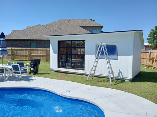 Elite Garage Door Services of Northwest ArkansasA backyard scene in Northwest Arkansas shows a white poolside shed with a newly installed black-framed overhead garage door. A ladder leans against the shed. Nearby, patio furniture and a blue swimming pool are visible under a clear sky.