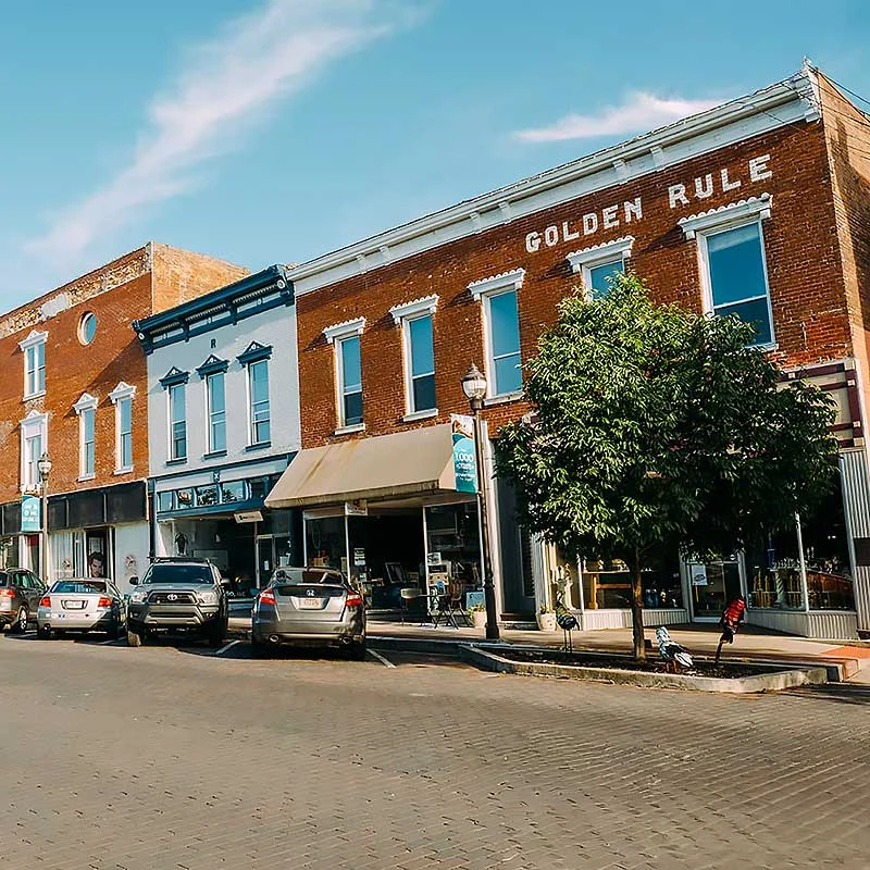 Elite Garage Door Services of Northwest Arkansas-A quaint street in Bentonville AR features historic brick buildings under a clear sky. Cars are parked along the street, with a small tree adding greenery. This local scene reflects the charm of Northwest Arkansas, ideal for residential garage doors.